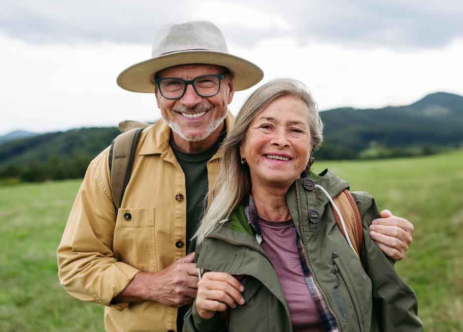 An older couple smiles joyfully together in a sunny field, surrounded by greenery and wildflowers.